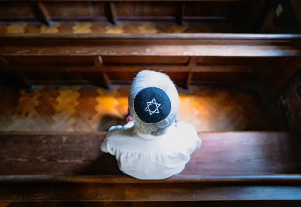 Close up image depicting a senior caucasian Jewish adult man in his 60s sitting inside a synagogue. He has his head bowed in prayer and he is wearing the traditional Jewish skull cap - otherwise known as a kippah or yarmulke - on his head. Horizontal color image with copy space.