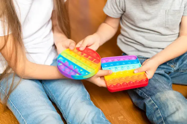 Photo of anti stress sensory pop it toy in a children's hands. a little happy kids sharing and plays with a simple dimple toys at home. toddlers holding and playing rainbow popit, trend 2021 year