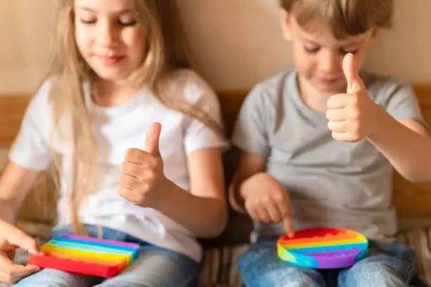 Photo of little happy kids plays with children's anti stress sensory pop it or simple dimple toys and showing hand like or thumb up. toddlers holding and playing popit rainbow bright color, trend 2021 year