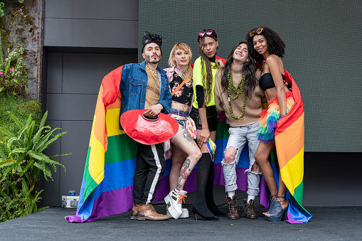 Group of friends of different sexual orientations celebrate gay pride wearing their rainbow flag and their multicolored outfits look towards the camera that is portraying them