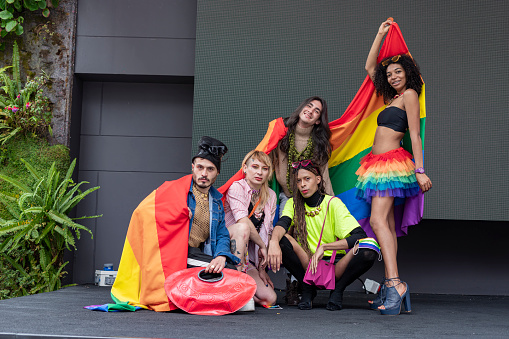Group of friends of different sexual orientations celebrate gay pride wearing their rainbow flag and their multicolored outfits look towards the camera that is portraying them