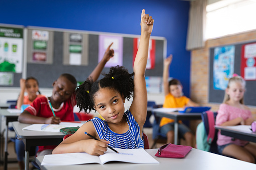 Niña afroamericana levantando las manos mientras está sentada en su escritorio en la clase en la escuela photo