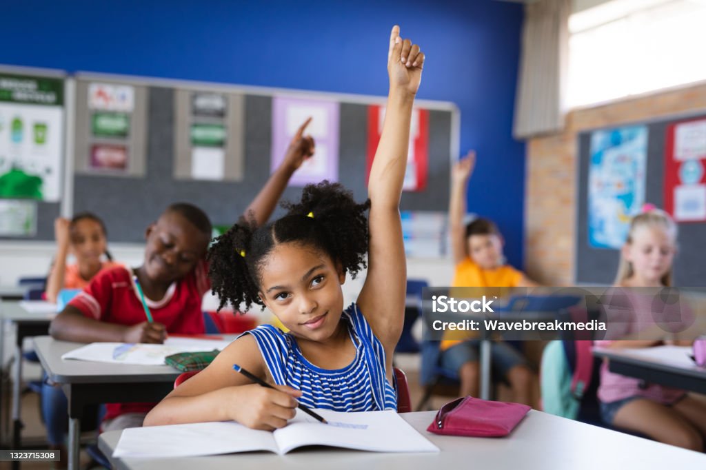 Menina afro-americana levantando as mãos enquanto estava sentada em sua mesa na classe na escola - Foto de stock de Criança royalty-free