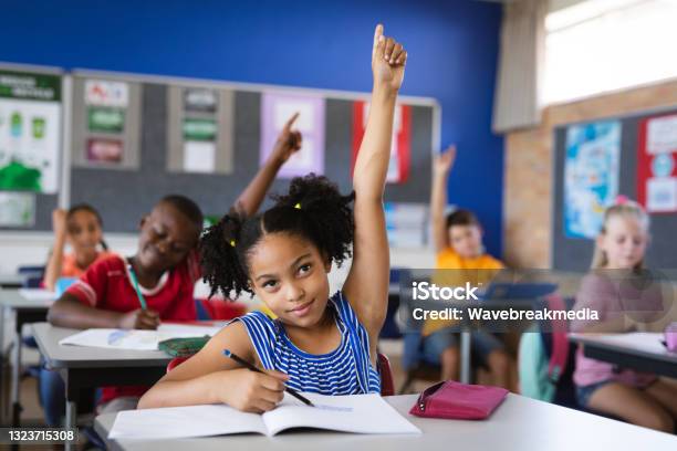 Foto de Menina Afroamericana Levantando As Mãos Enquanto Estava Sentada Em Sua Mesa Na Classe Na Escola e mais fotos de stock de Criança