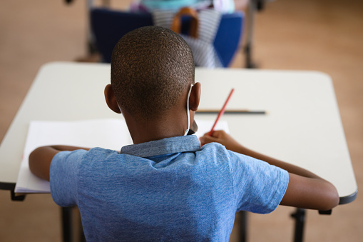 Rear view of african american boy studying while sitting on his desk in the class at school. education back to school health safety during covid19 coronavirus pandemic