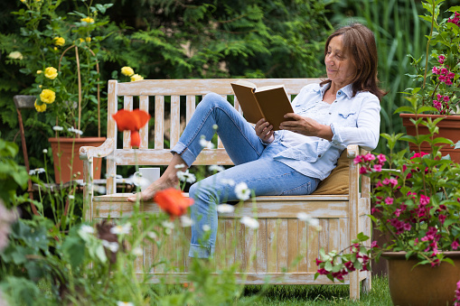 Smiling senior woman wearing orange blouse and straw hat sitting on bench in garden and using smart phone.