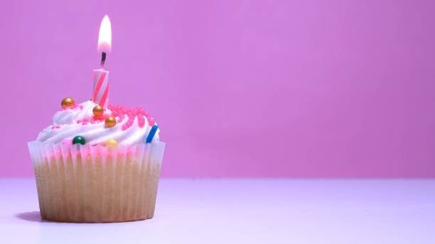 istock Cupcake with burning festive candle at white cream and sugar icing on pink background, macro shallow depth of field view. Homemade vanilla cup cake with buttercream frosting close up. 1323716099