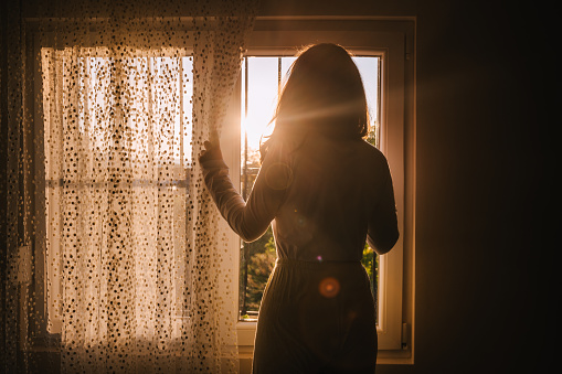 A young adult woman looking through a window with the bars on moving the curtain
