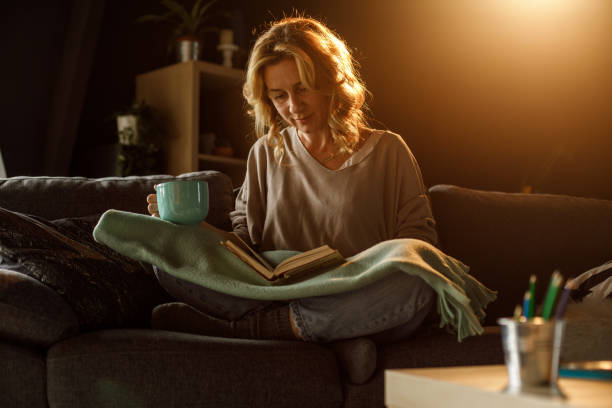 istock Smiling woman reading a book and enjoying tea on the sofa 1323730697