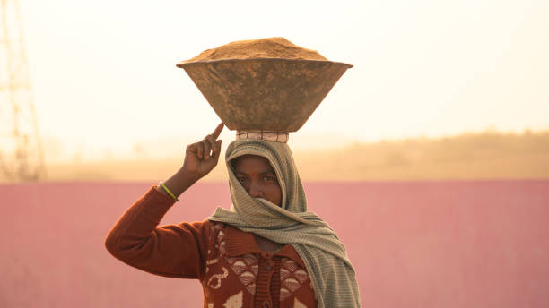 travailleuse indienne tenant un contenant de matières premières sur la tête pendant les travaux sur le chantier de construction - porter sur la tête photos et images de collection