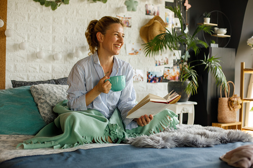 Copy space shot of cheerful mid adult woman sitting in bed in the morning, relaxing, enjoying a cup of coffee and contemplating while reading a book.