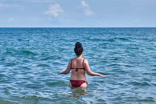 A young asian woman with hairstyle from dreadlocks on black hair, in a red swimsuit, walking into the sea. View from the back. Summer seascape.