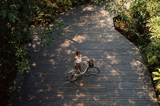 Beautiful happy young Caucasian woman exploring a tropical public park by bicycle on a sunny day She is standing on the wooden pathway holding the bicycle and looking around