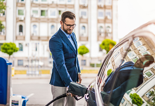 Young businessman sipping fuel into his car tank at the gas station.