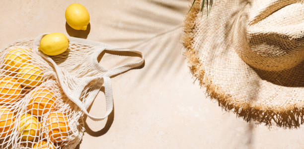 Summer flat lay banner on beige background. Straw hat and lemon fruits in eco friendly mesh shopping bag. Trendy palm shadow and sunlight, sun. Minimal summer travel fashion concept. Top view stock photo
