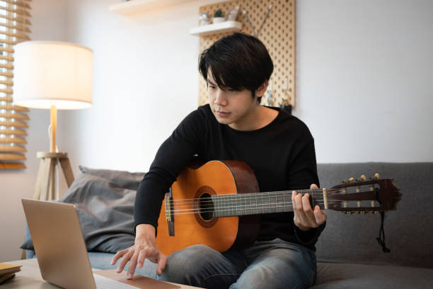 istock Young man using laptop and playing guitar in living room. 1323803492