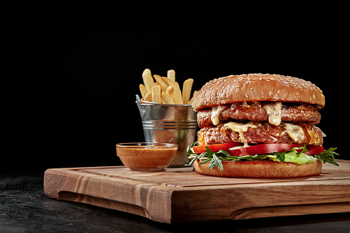Closeup of appetizing double hamburger with two juicy patties, caramelized onion, fresh tomato and greens in fluffy sesame seed bun served on wooden board with fries and sauce. Fast food concept