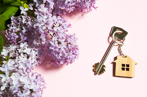 House key with keychain on pink spring background and lilac branches. Summer holiday home, booking cottage in the countryside, moving to new home, mortgage, rent and purchase real estate. Copy space