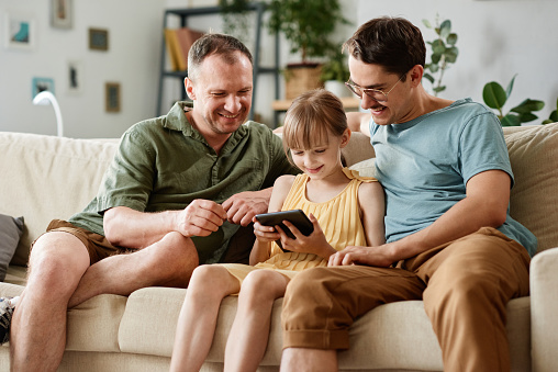 Gay male couple sitting on sofa with little girl while she playing on digital tablet in the room