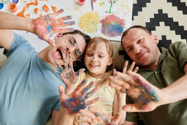 Gay family painting with daughter High angle view of happy gay family lying on the floor with their daughter showing their painted hands and smiling they painting at home gay person stock pictures, royalty-free photos & images
