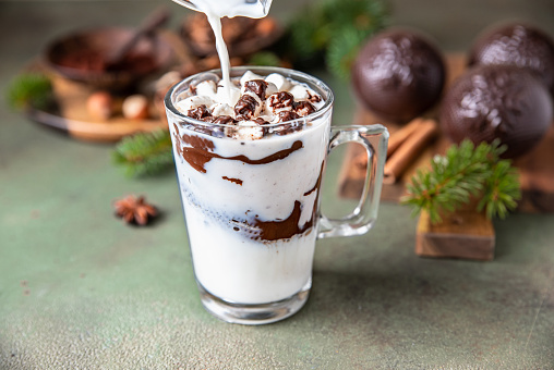 Hot chocolate with marshmallow and cocoa or chocolate bomb on green concrete background. Winter composition with fir branches and spices. Selective focus.