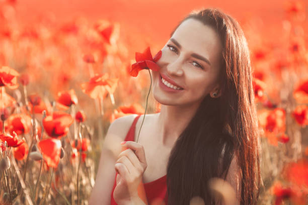 jeune femme brune 25-30 ans avec de longs cheveux soignés en robe rouge profiter d’une fleurs de pavot dans le champ - cosmetics young women human skin 25 30 years photos et images de collection