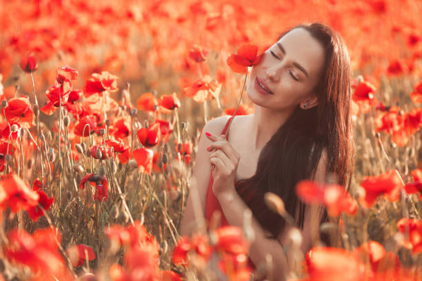 jeune femme brune 25-30 ans avec de longs cheveux soignés en robe rouge profiter d’une fleurs de pavot dans le champ - cosmetics young women human skin 25 30 years photos et images de collection