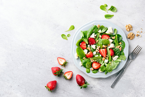 Summer strawberry salad with spinach, walnuts and goat cheese served on a plate, top view, grey concrete background, copy space for text or design