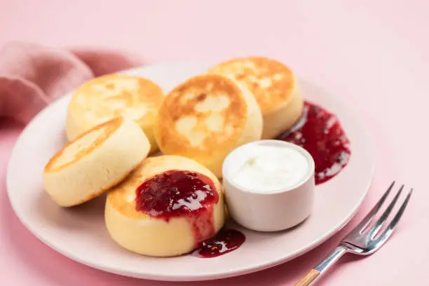 Syrniki, cottage cheese fritters with raspberry jam and sour cream on a pink plate, closeup view