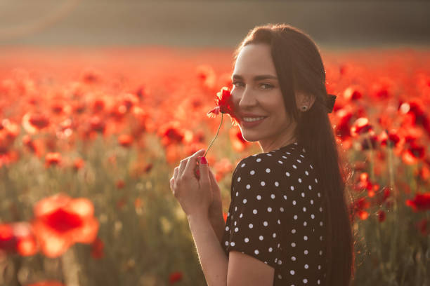 jeune femme brune 25-30 ans avec de longs cheveux soignés en robe noire pois blancs profiter d’une fleur de pavot dans le champ - cosmetics young women human skin 25 30 years photos et images de collection