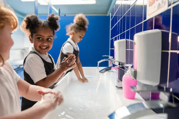 A side-view of diverse UK young schoolgirls, going through COVID-protocols and washing their hands with soap and water in their school toilets. They are all wearing their school uniforms.