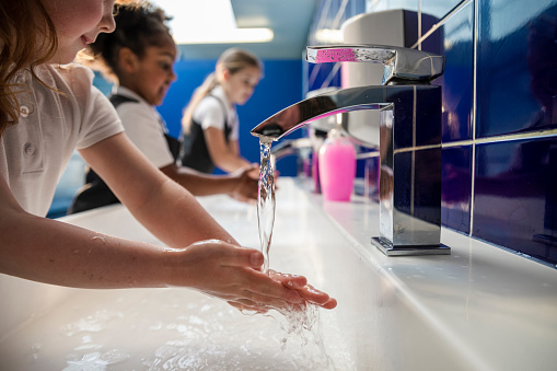A side-view of diverse UK young schoolgirls, going through COVID-protocols and washing their hands with soap and water in their school toilets. They are all wearing their school uniforms.