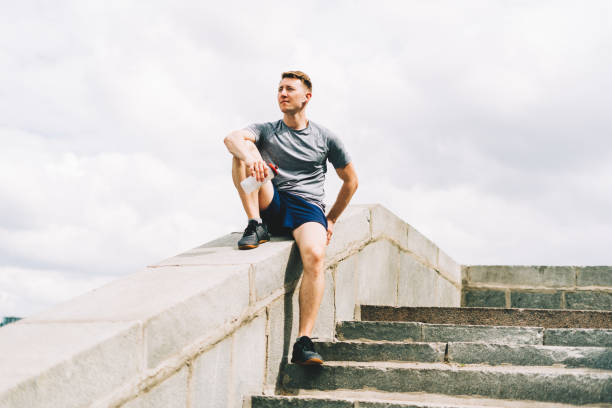 istock Tired Young man runner sitting and relaxing after sport training. Holding water bottle while doing workout in summer city street 1323843167
