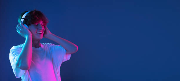 Caucasian young man's portrait on dark studio background in neon. Concept of human emotions, facial expression. Listening to music. Caucasian young man's portrait on dark studio background in neon light. Handsome male model in casual style. Concept of human emotions, facial expression, youth. Copy space for ad. neon portrait boy stock pictures, royalty-free photos & images