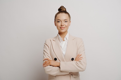 Picture of young beautiful woman manager in beige suit and white shirt with crossed arms, slightly smiling and looking straight forward while being succesfull and confident against to light wall