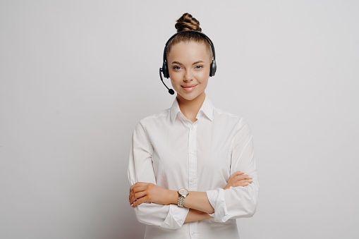 Confident female air traffic controller in shirt wearing black headset, standing with crossed arms communicating with plane pilots, standing against light wall. Portrait of call center worker