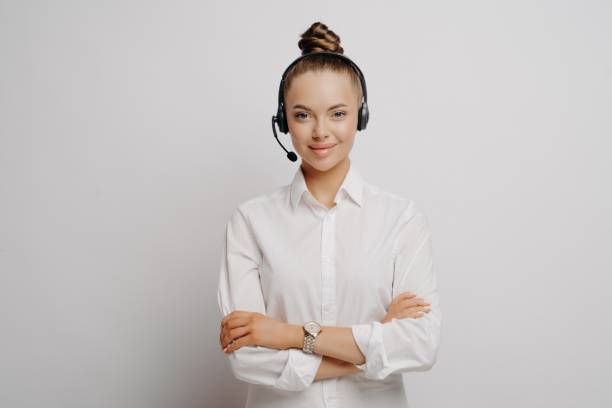 Confident female air traffic controller in white shirt Confident female air traffic controller in shirt wearing black headset, standing with crossed arms communicating with plane pilots, standing against light wall. Portrait of call center worker telephone worker stock pictures, royalty-free photos & images