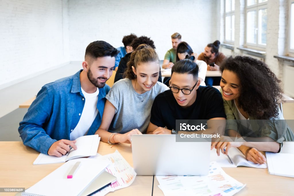 Classmates are discussing together in classroom Young man reading on laptop while sitting by male and female student at desk. Classmates are discussing together in classroom. They are doing assignment. 20-24 Years Stock Photo