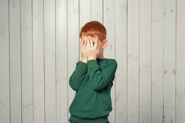 niño pequeño de jengibre asustado escondiendo la cara en las palmas de las manos y mirando hacia fuera con un ojo entre sus dedos - avergonzamiento fotografías e imágenes de stock