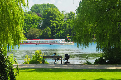 Hamburg, Germany - July, 2019: Alster lake and Alsterpark in Hamburg, Germany. Aussenalster lake, place for walking. Pleasure boat with tourists, a grandmother with a grandson sits on a bench