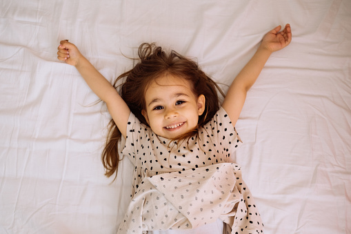 Carefree little kid girl sleeping in his white bed . Funny happy child playing and smiling. Family, vacation, childhood concept