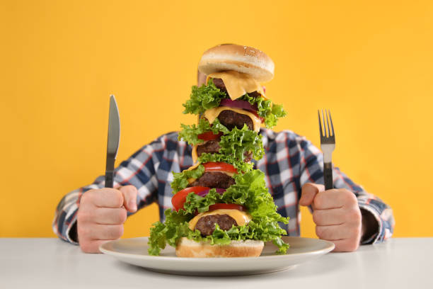 Hungry man with cutlery and huge burger at white table on yellow background Hungry man with cutlery and huge burger at white table on yellow background big plate of food stock pictures, royalty-free photos & images