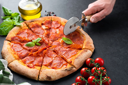 Hand slicing pepperoni pizza on black concrete background. Tasty hot italian pizza cut into slices