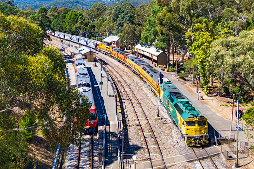Belair, Australia - April 04, 2021: Aerial view One Rail Australia loaded grain train with five (5) diesel locomotives passing the historic Belair Railway Station in the Adelaide Hills on its way to unload at the Viterra terminal in Port Adelaide. Waiting at the station platform at left is an Adelaide Metro diesel commuter train.