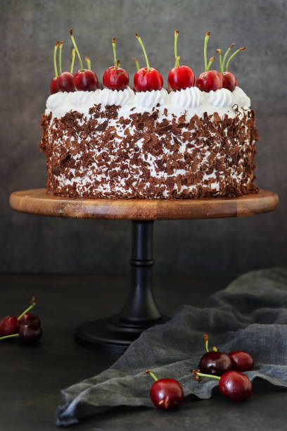 istock Image of Black Forest gateau with piped whipped cream rosettes topped with morello cherries, luxury chocolate cake frosted with whipped cream and covered with chocolate shavings on cake stand, black background, focus on foreground 1323862458