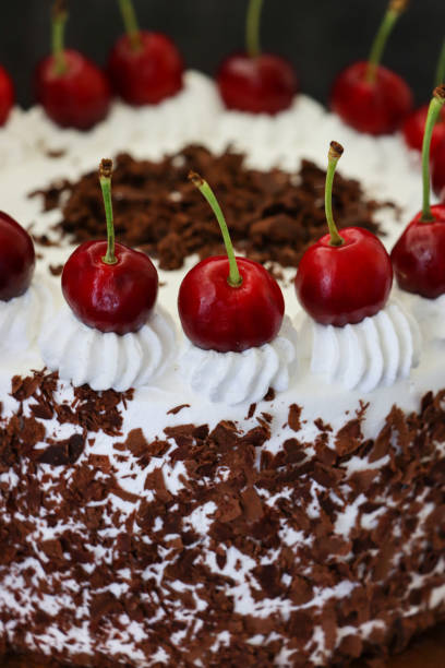 istock Image of Black Forest gateau with piped whipped cream rosettes topped with morello cherries, luxury chocolate cake frosted with whipped cream and covered with chocolate shavings on cake stand, black background, elevated view, focus on foreground 1323862476