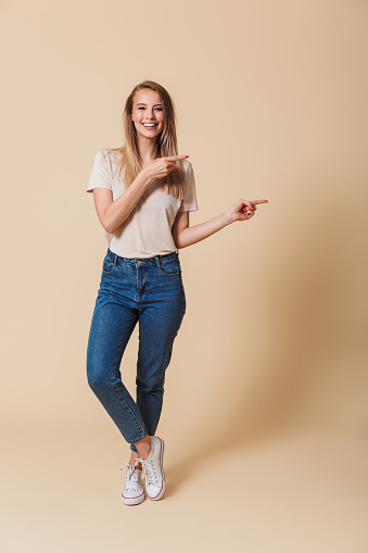 Full length portrait of a happy casual girl pointing finger away at copy space isolated over beige background