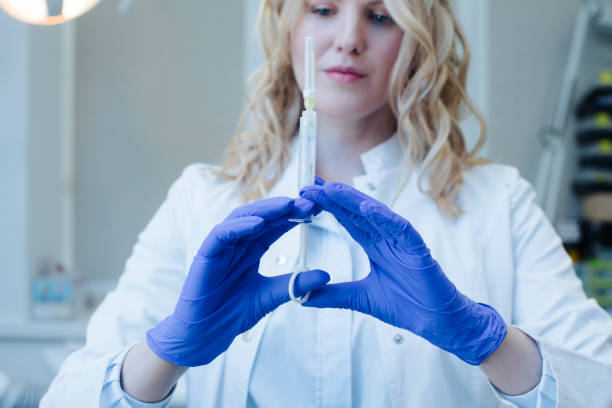 Blonde doctor woman in a white coat and blue medical gloves, holding a syringe with an anesthetic in a solution in front of him. close-up, focus on hands stock photo