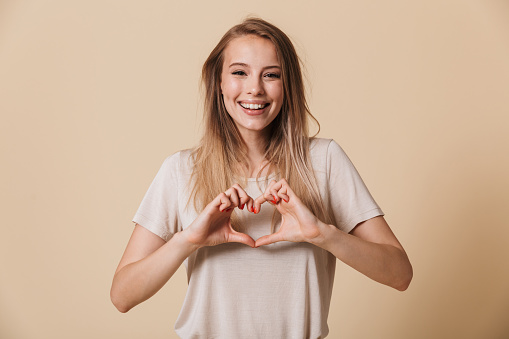 Portrait of a lovely young woman showing love gesture with her fingers isolated over beige background