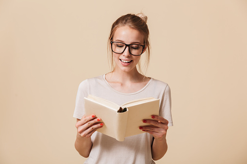 Portrait of a smiling young girl in eyewear reading a book isolated over beige background